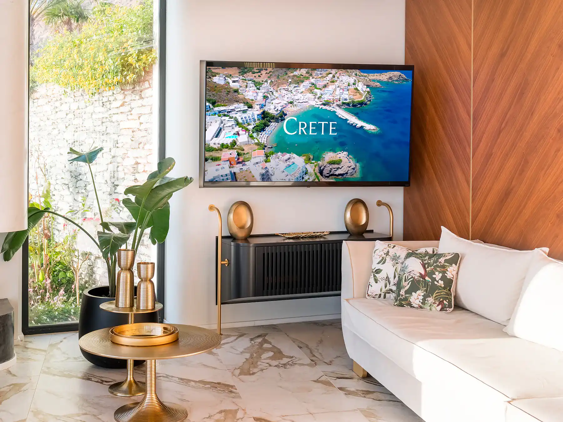 Bright living room with white sofas, golden accents, and Cretan landscape visible through large windows.