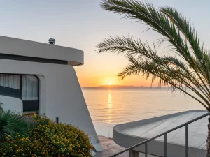 Evening view from villa terrace with palm tree and sun setting over the Aegean Sea.